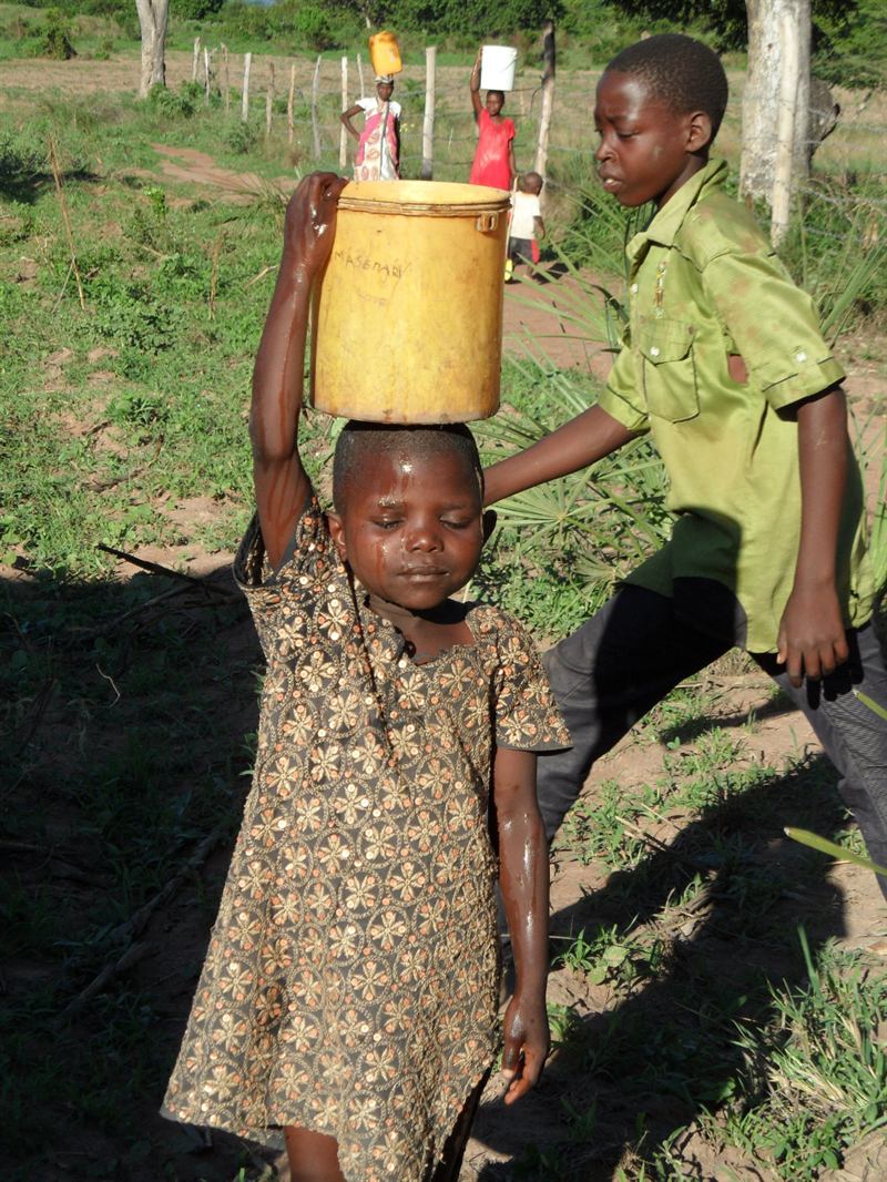 A young girl carries a heavy water container on her head along a rural path